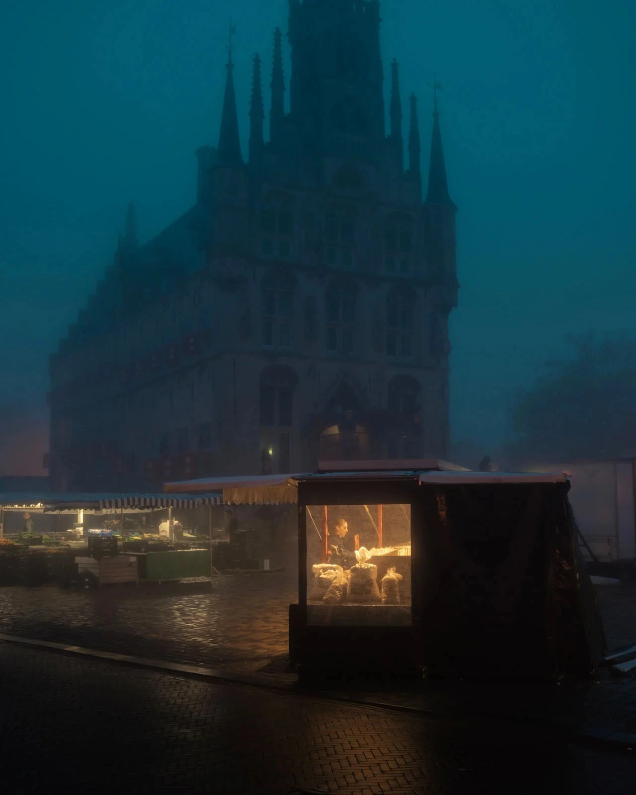Toplo osvijetljeni tržni štand na mokroj kaldrmi ispred gotičkog zdanja u plavoj večernjoj magli, serija Morning Ritual, pobjednik kategorije ulične fotografije